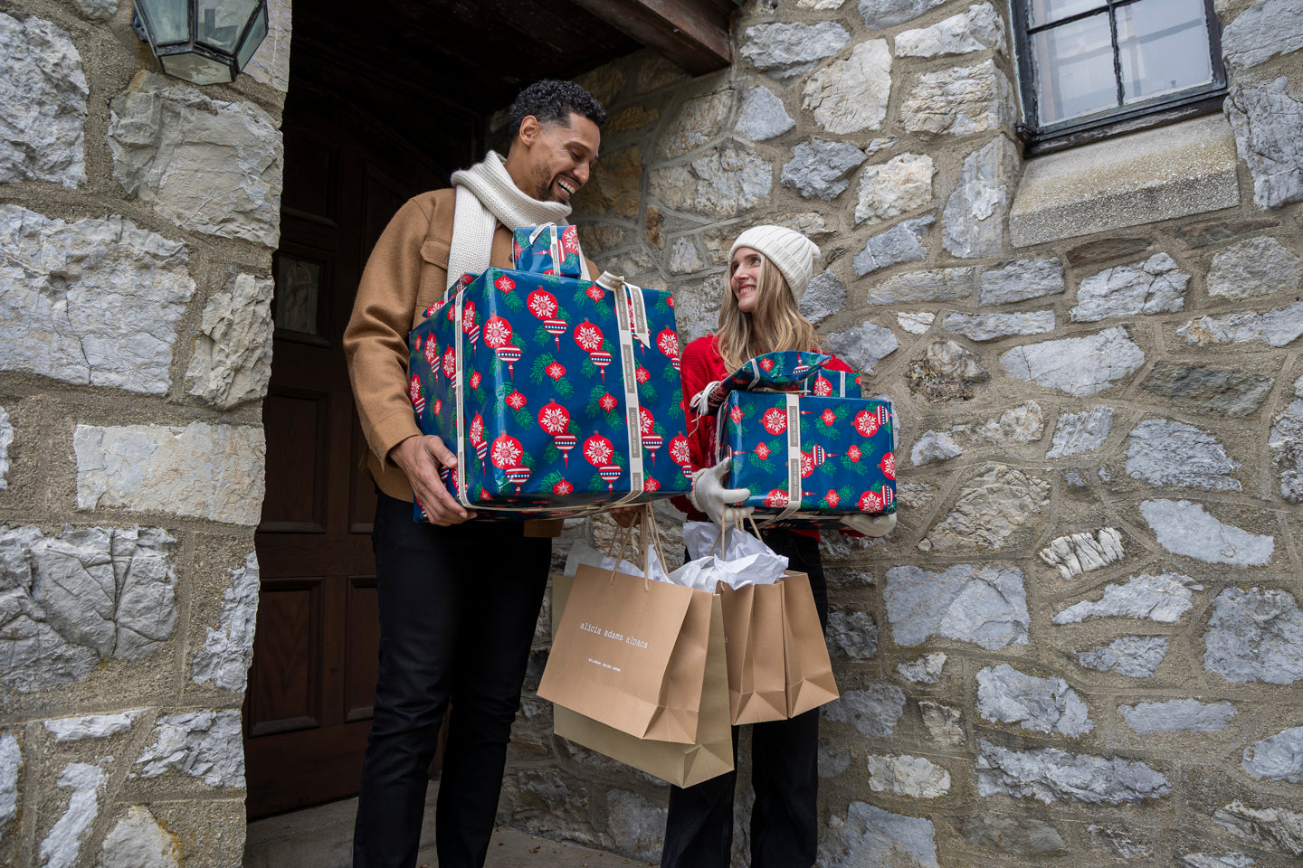 Couple holding colorful gifts at a stone house entrance.